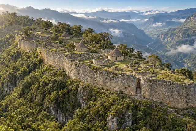 Muraille de pierre d’une forteresse sur une crête boisée, maisons rondes dont trois couvertes de chaume, passage étroit dans le mur, et vallée profonde dans les nuages