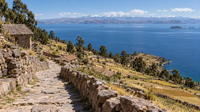 Chemin de pierre entre des murets sur une île, champs en terrasses et lac bleu immense, sommets enneigés sur l’autre rive